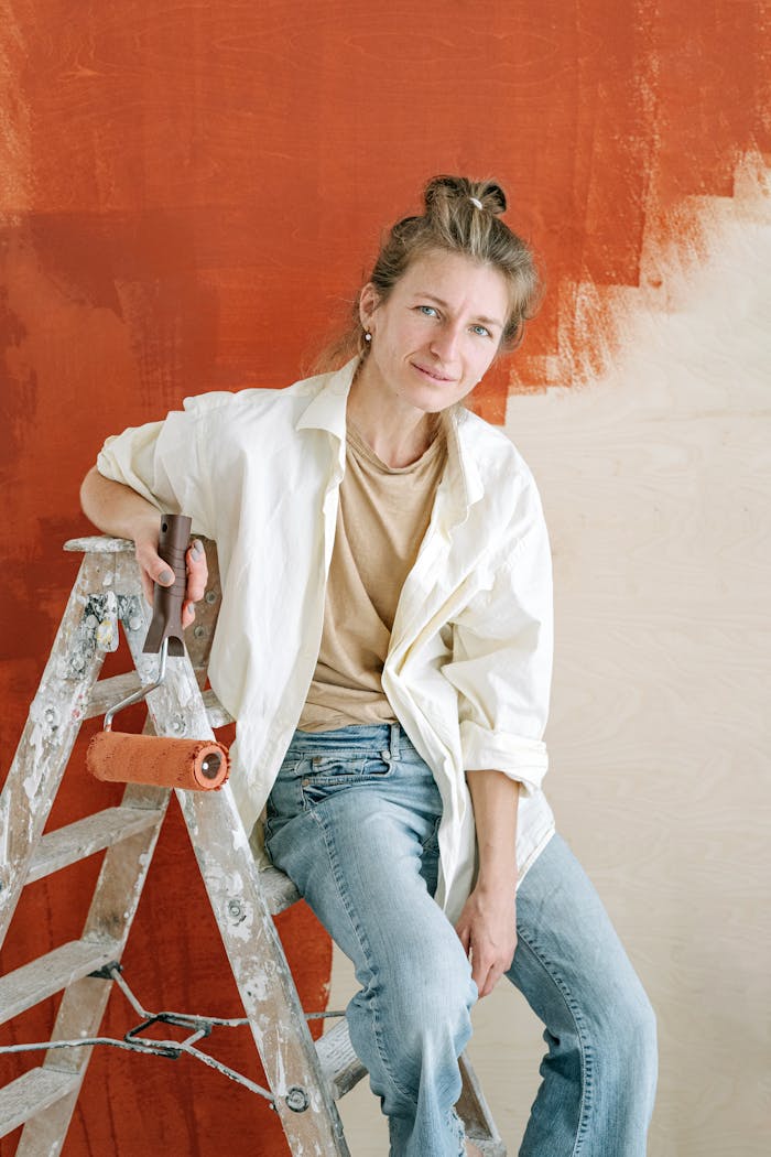 Woman sitting on a ladder with a paint roller, smiling during home renovation.